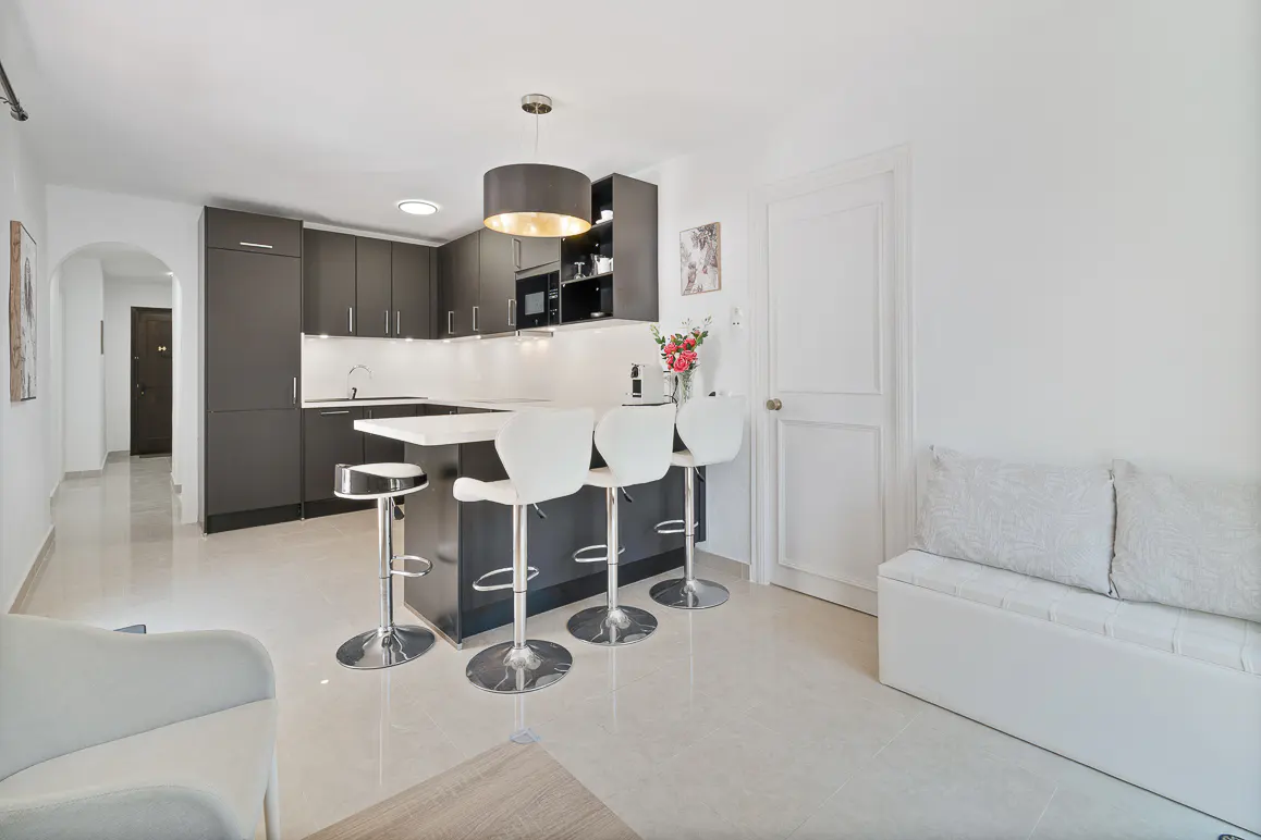 A modern kitchen with dark cabinets, a white countertop island with three white bar stools, and a white bench.