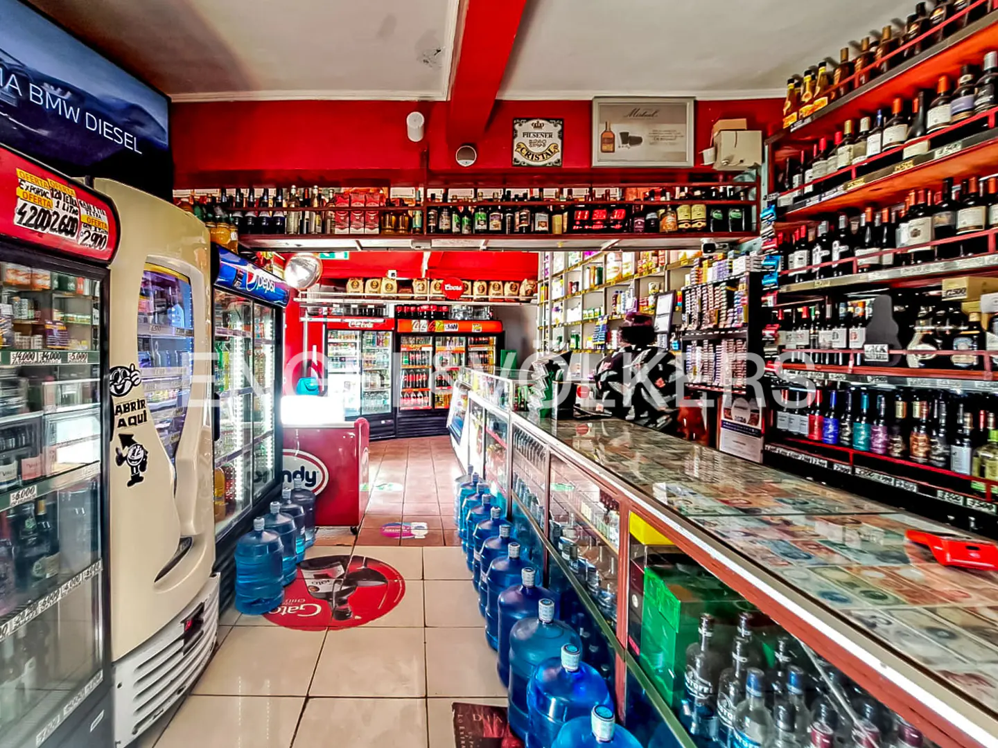 Interior view of a small grocery store with red walls, shelves stocked with bottles, and refrigerators filled with drinks.