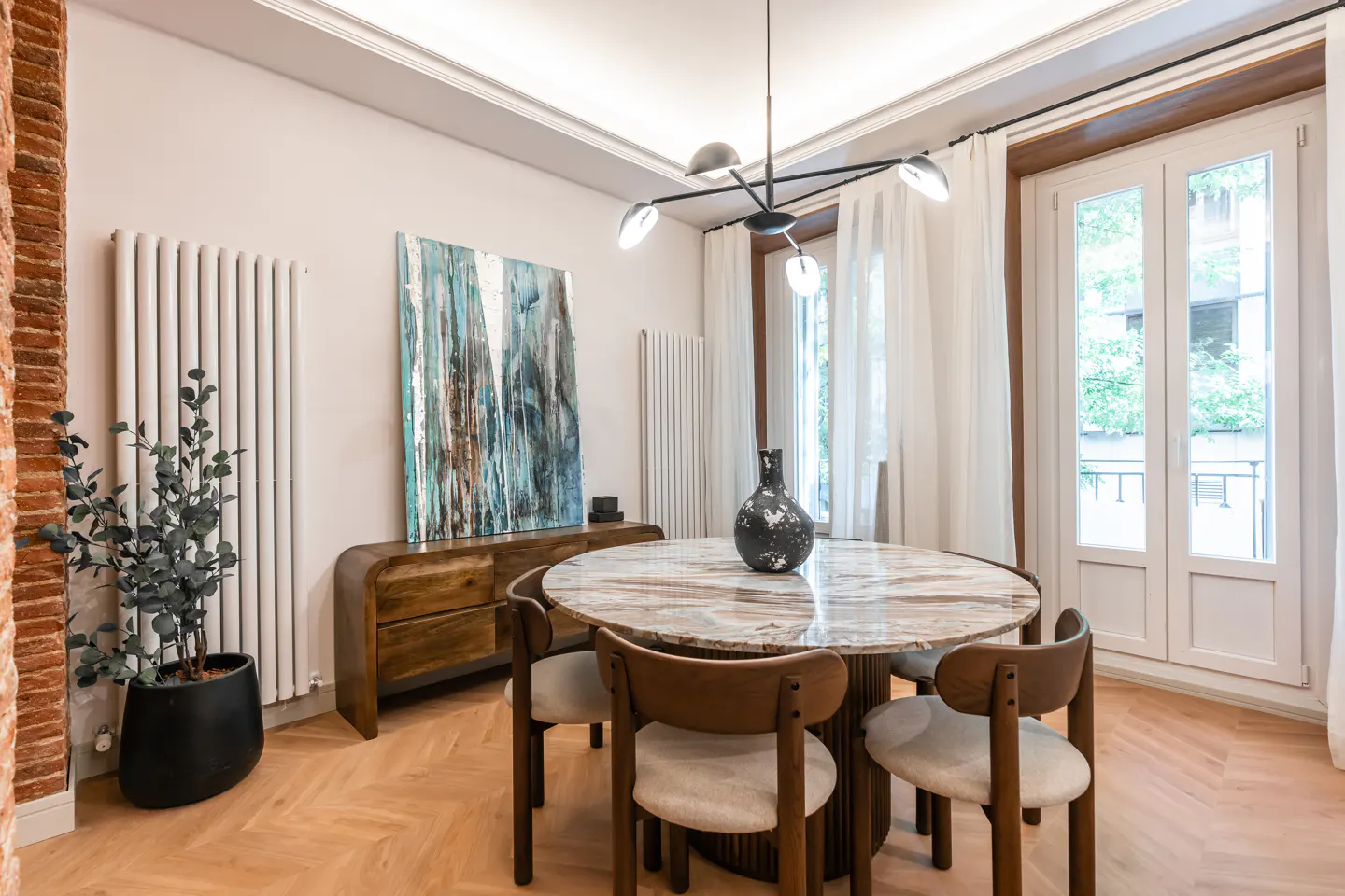 A dining room with a round marble table, wooden chairs, and a modern chandelier. A blue abstract painting hangs above a wooden sideboard. White walls and herringbone wood floors.