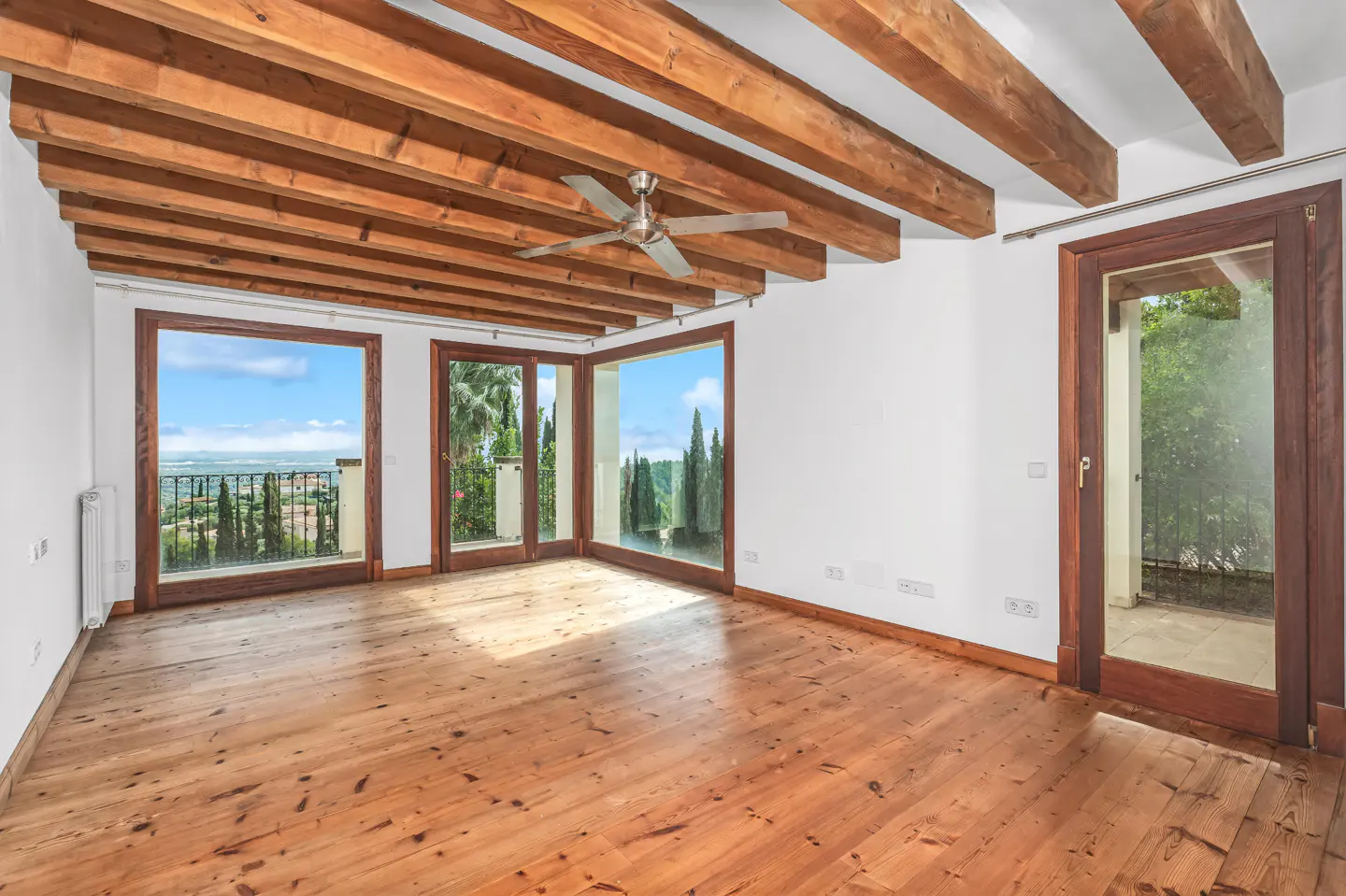 Bright room with wood floors, white walls, and exposed wood beam ceiling. Large windows offer views of greenery and a distant landscape.