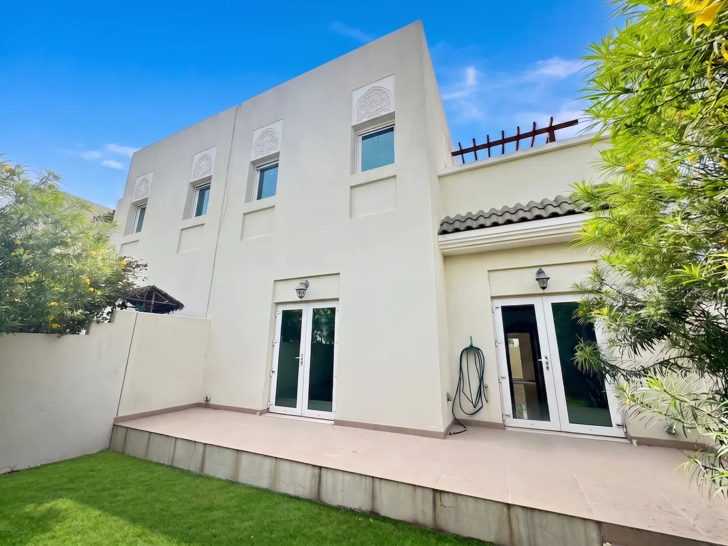 Exterior view of a two-story, off-white house with a green lawn and blue sky. French doors open to a patio.
