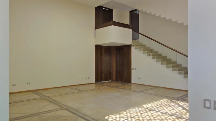 An interior view of a room with a staircase, wooden door, and patterned floor. The walls are off-white, and sunlight streams in.