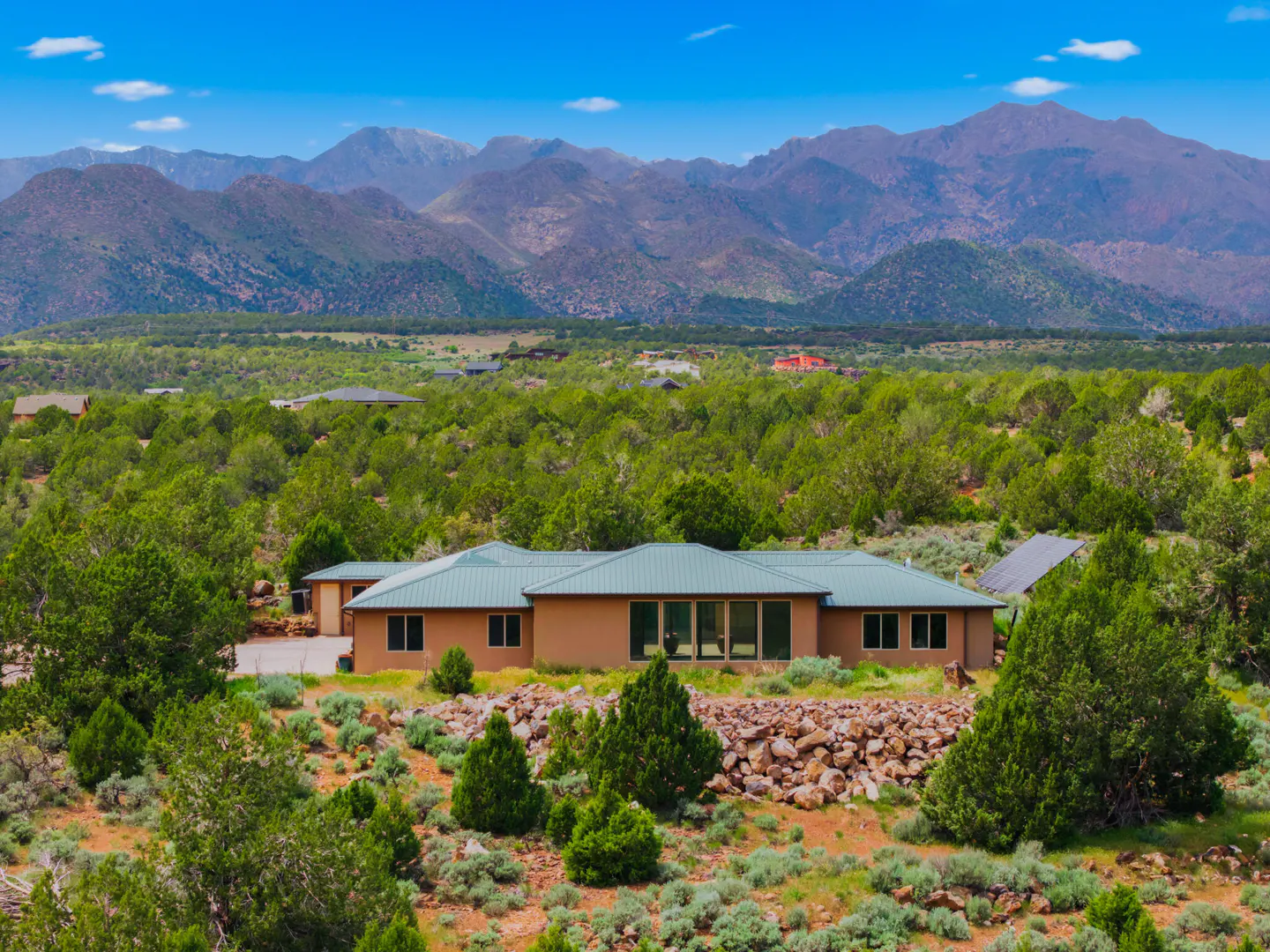 Tan house with a green roof surrounded by trees, with mountains in the background under a blue sky.