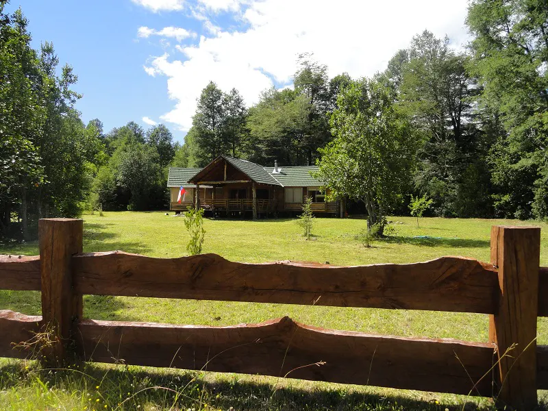 A log cabin with a green roof sits on a grassy lawn, framed by a rustic wooden fence and lush green trees.