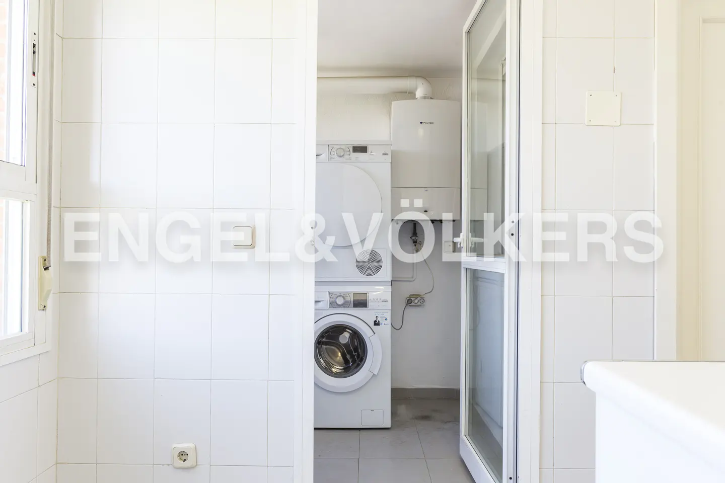 A bright laundry room with white tiled walls, a stacked washer and dryer, and a white-framed glass door.