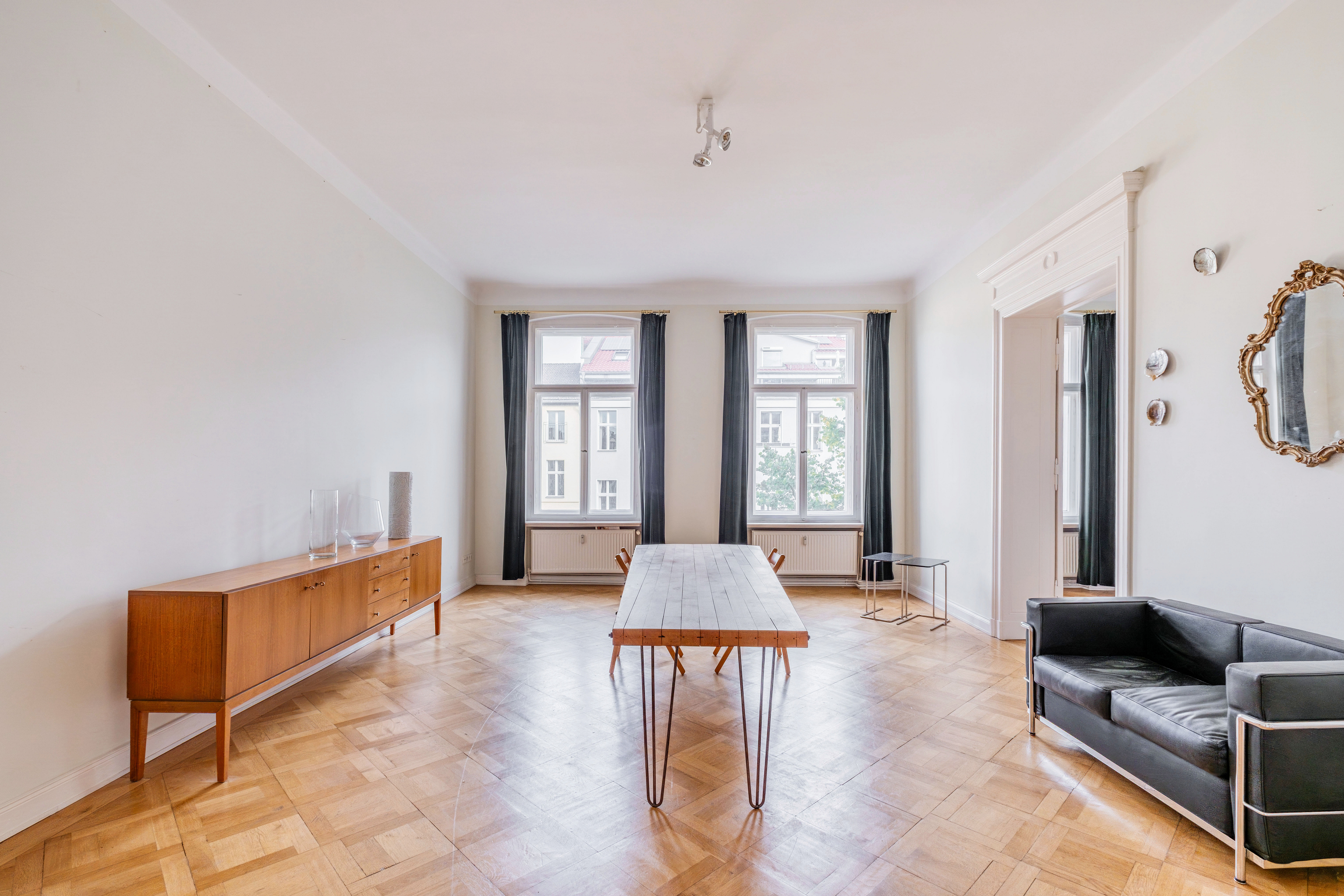 Spacious room with parquet floors, a wooden sideboard, a central dining table with hairpin legs, and a black leather sofa. [AI-generated text]