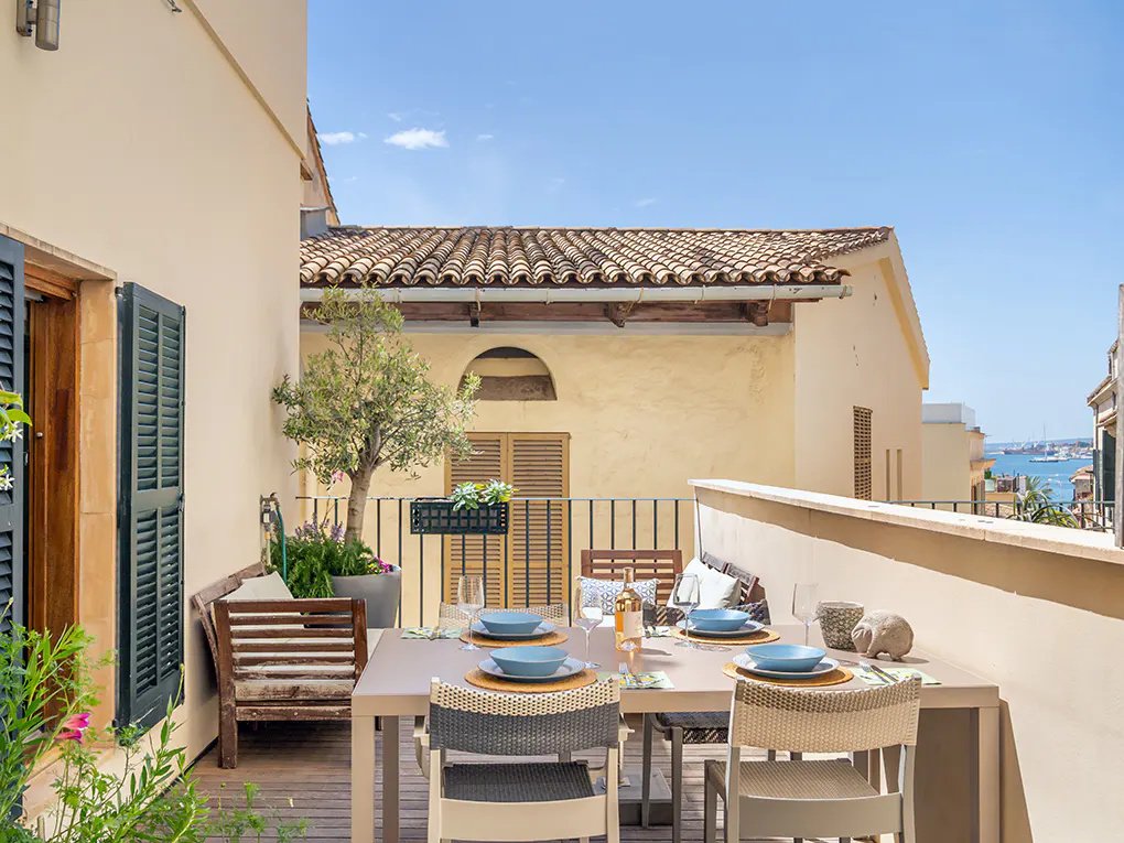 Outdoor dining area on a balcony with a table set for a meal, chairs, and a view of buildings and the sea.