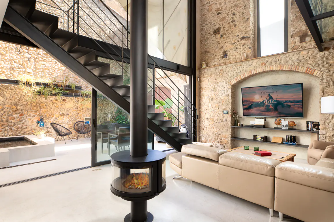 Interior view of a modern living room with stone walls, a black fireplace, and a beige sofa. Black metal stairs lead to the upper level. A courtyard is visible through glass doors.