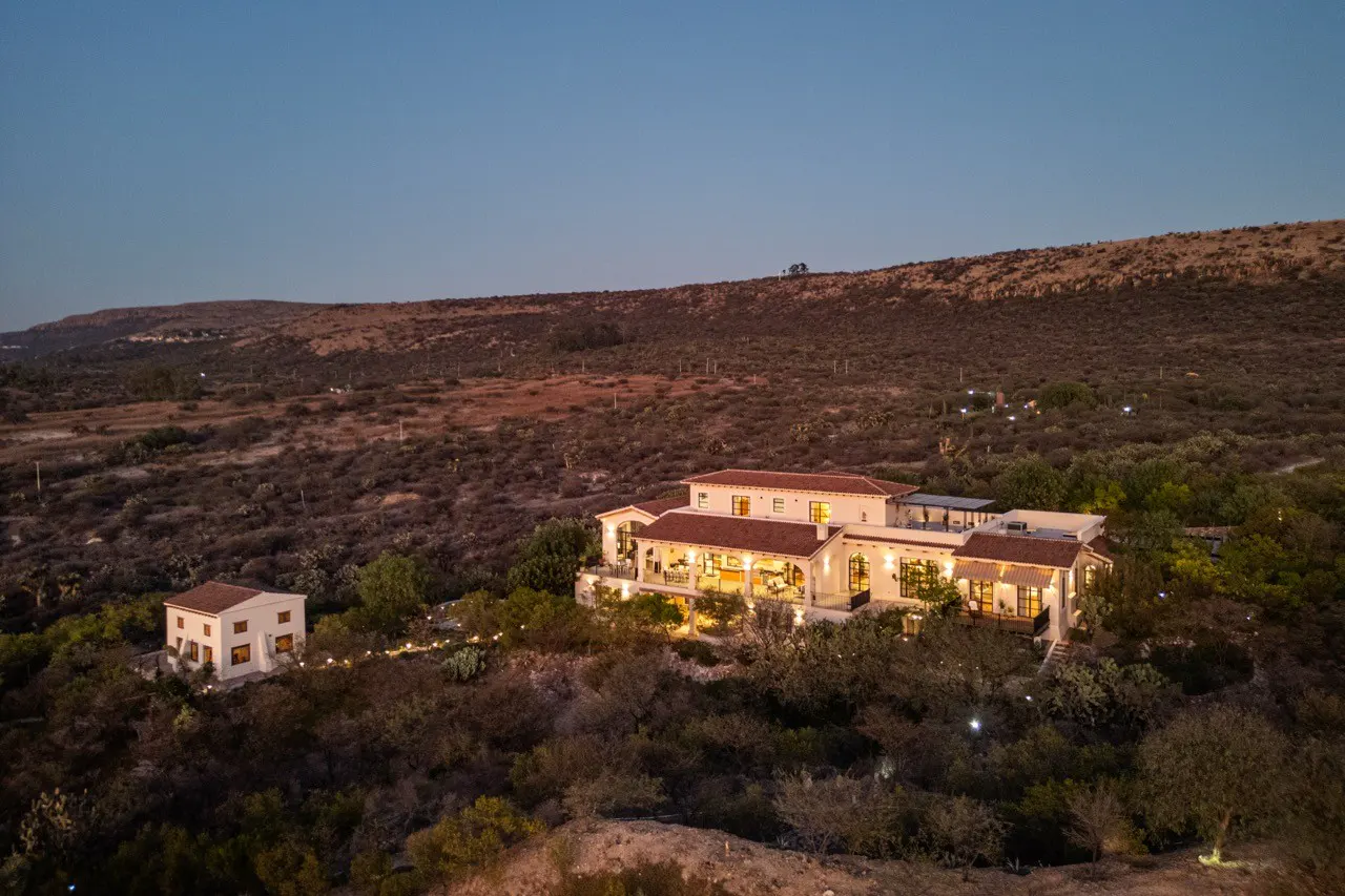 A tan villa with a red tile roof sits on a hill surrounded by trees and desert landscape at dusk.