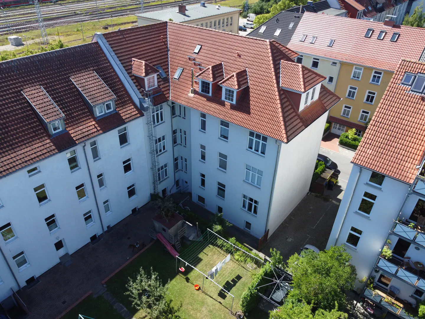 Aerial view of a white apartment building with a red tile roof and a green backyard with a swing set.