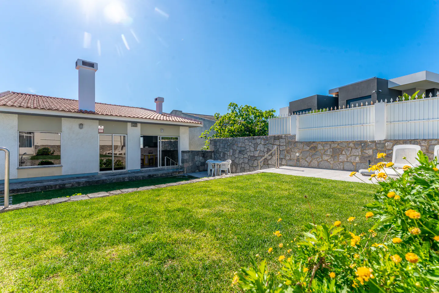 A sunny backyard with green grass, yellow flowers, and a white house with a red tile roof. A stone wall and white fence are in the background.