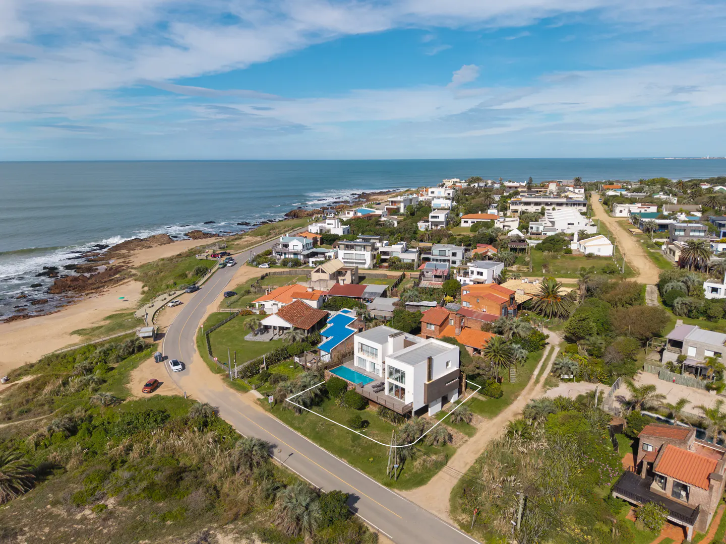Aerial view of a modern white house with a pool near the ocean and other houses in a coastal community.