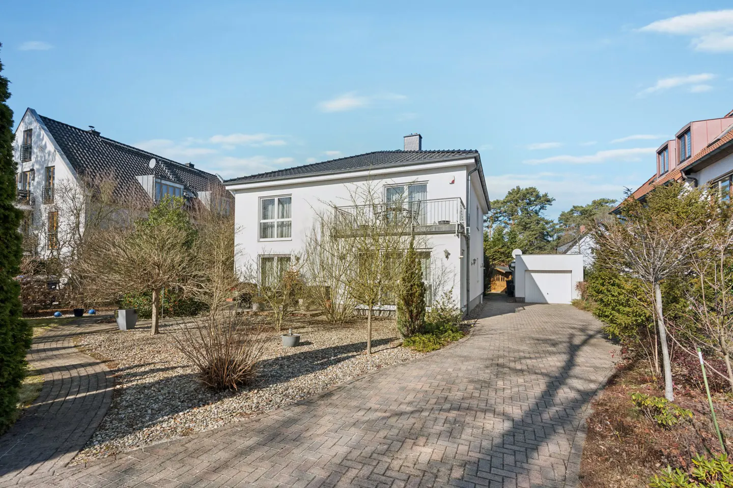 Two-story white house with a black roof and a balcony, surrounded by trees and a brick driveway.