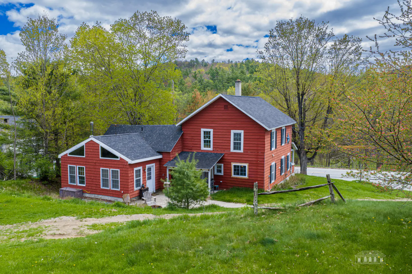 Two-story red farmhouse with a gray roof, surrounded by green trees and grass, under a cloudy sky.