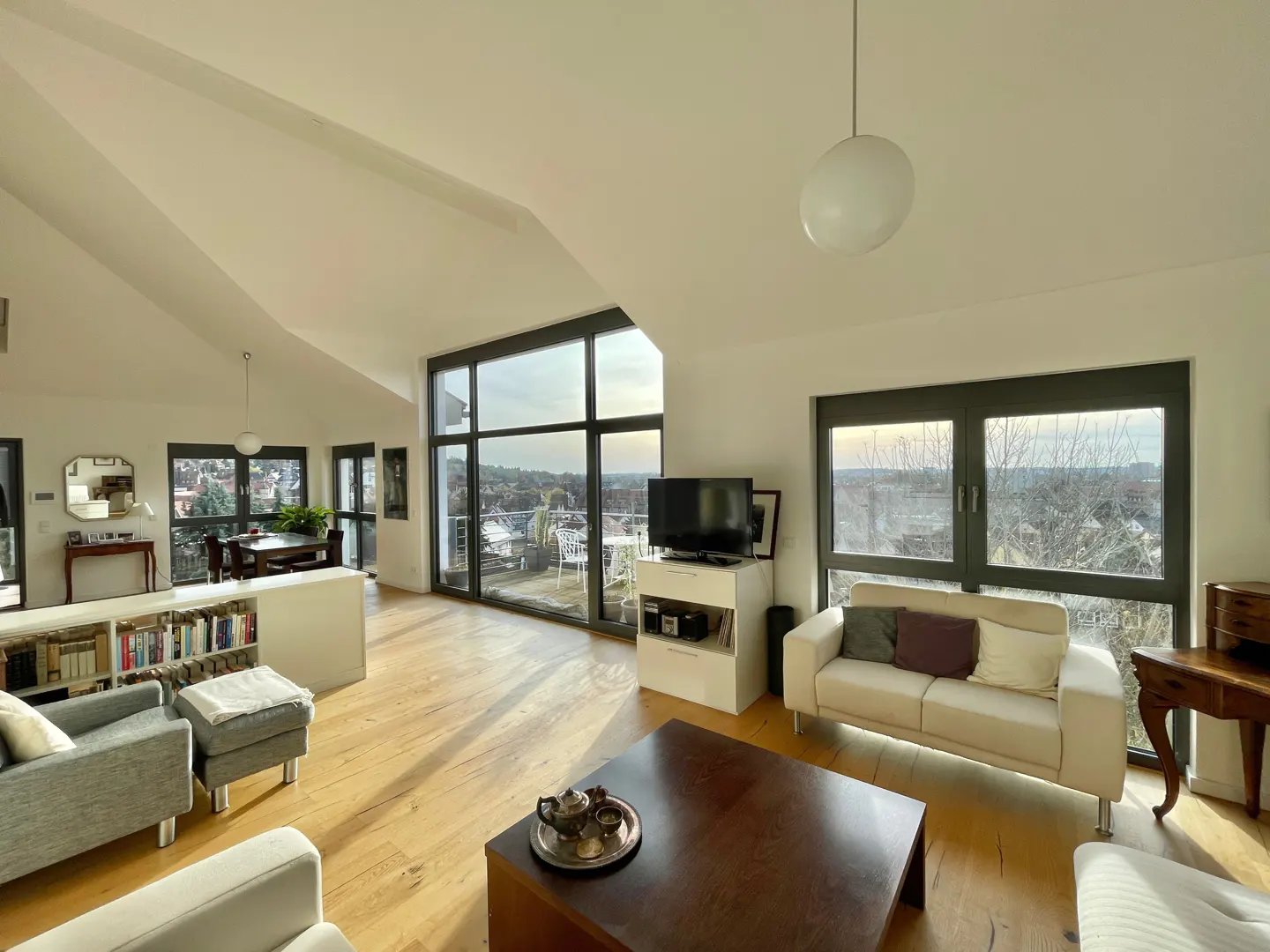 Bright living room with wood floors, white walls, and large windows. A gray sofa and ottoman are in the foreground, with a dark wood coffee table. A white sofa sits near a window with a view.