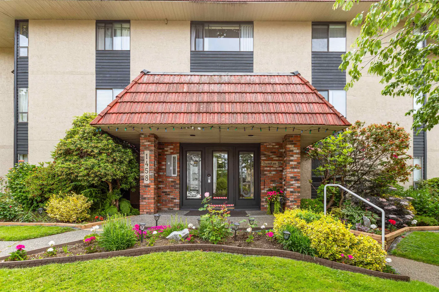 Apartment building entrance with red tile roof, brick pillars, and black double doors. Manicured lawn and flower beds in front.