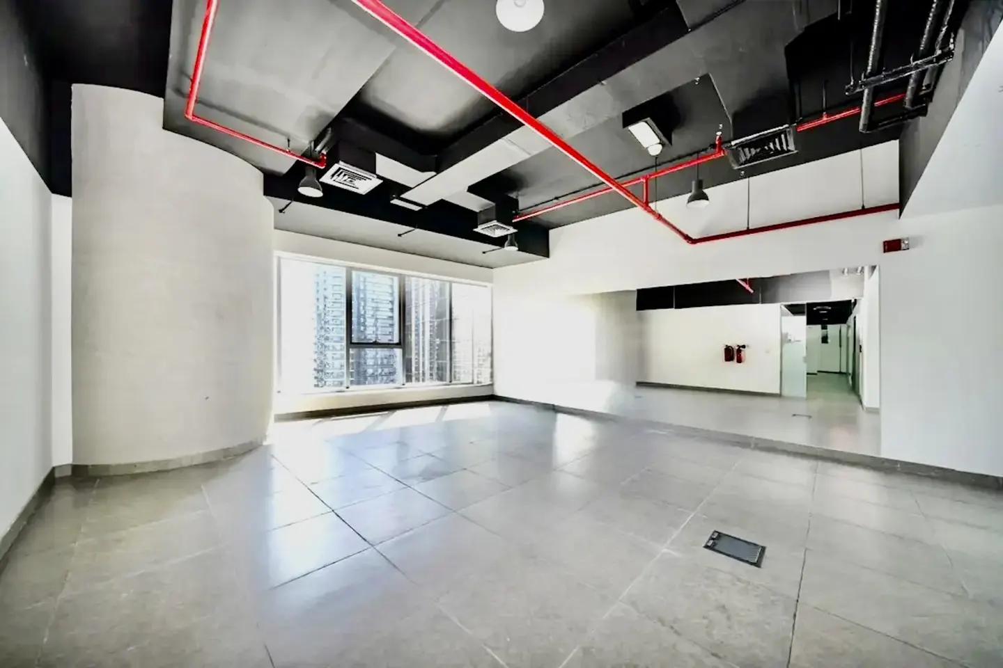 Empty office space with gray tile floor, white walls, and black ceiling with red pipes. Large windows overlook city buildings.
