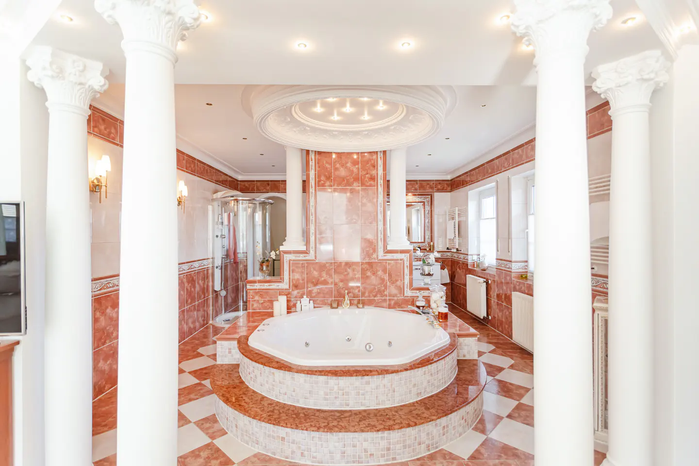 Opulent bathroom with white columns, red-and-white checkered floor, and a raised, round jacuzzi tub. Shower and vanity visible.