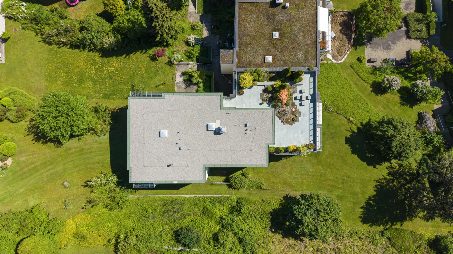 Aerial view of a modern home with a flat roof, a stone patio, and lush green landscaping surrounding the property.