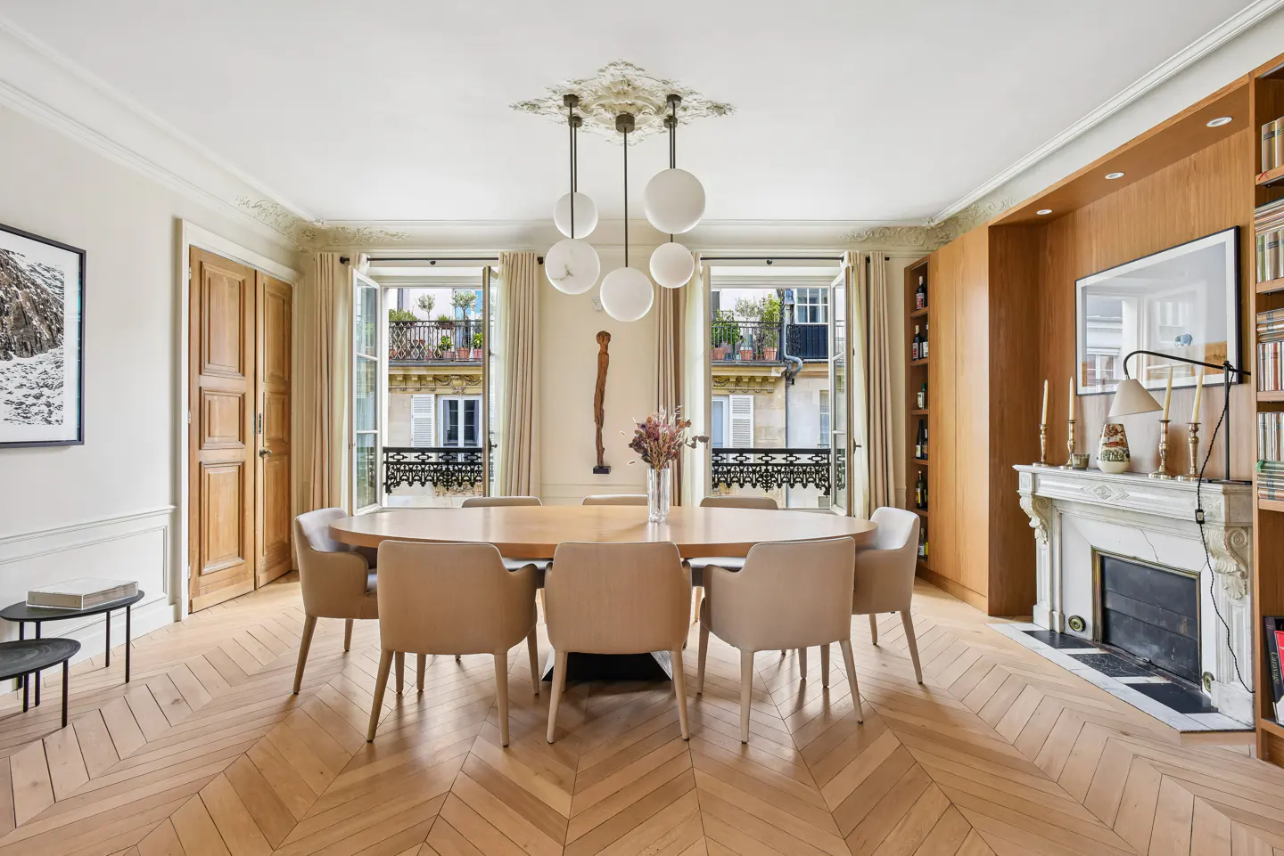 Bright dining room with herringbone wood floors, oval table with beige chairs, and open windows overlooking a Parisian street.