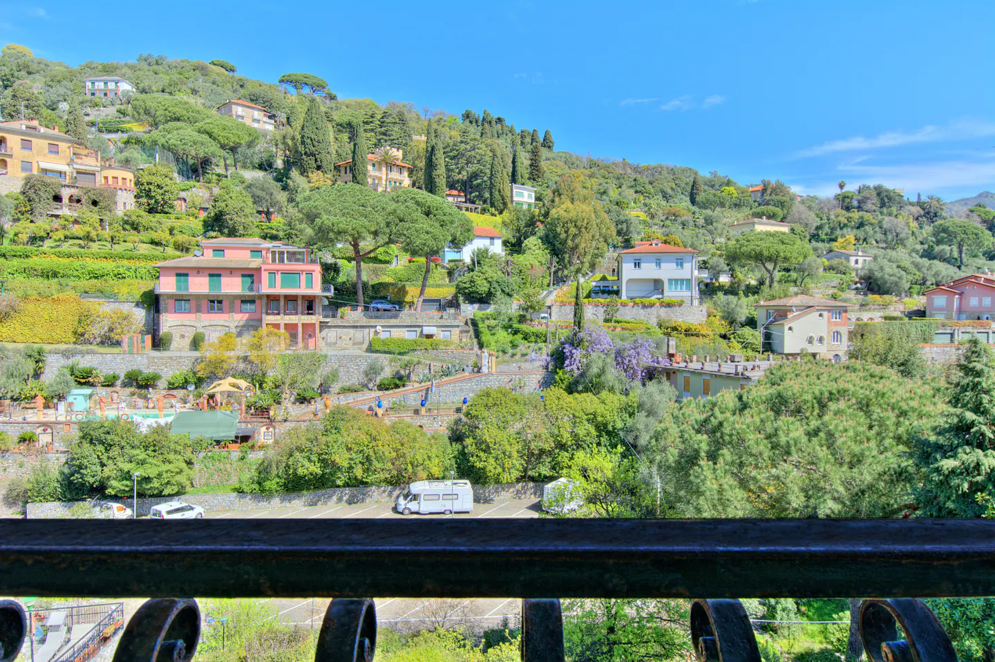 View from a balcony of a hillside with houses, trees, and a parking lot with cars.