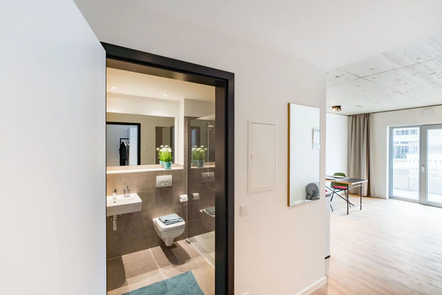 A modern apartment hallway with a bathroom view. The bathroom has a sink, toilet, and mirror. The hallway leads to a living area with a table and chairs.