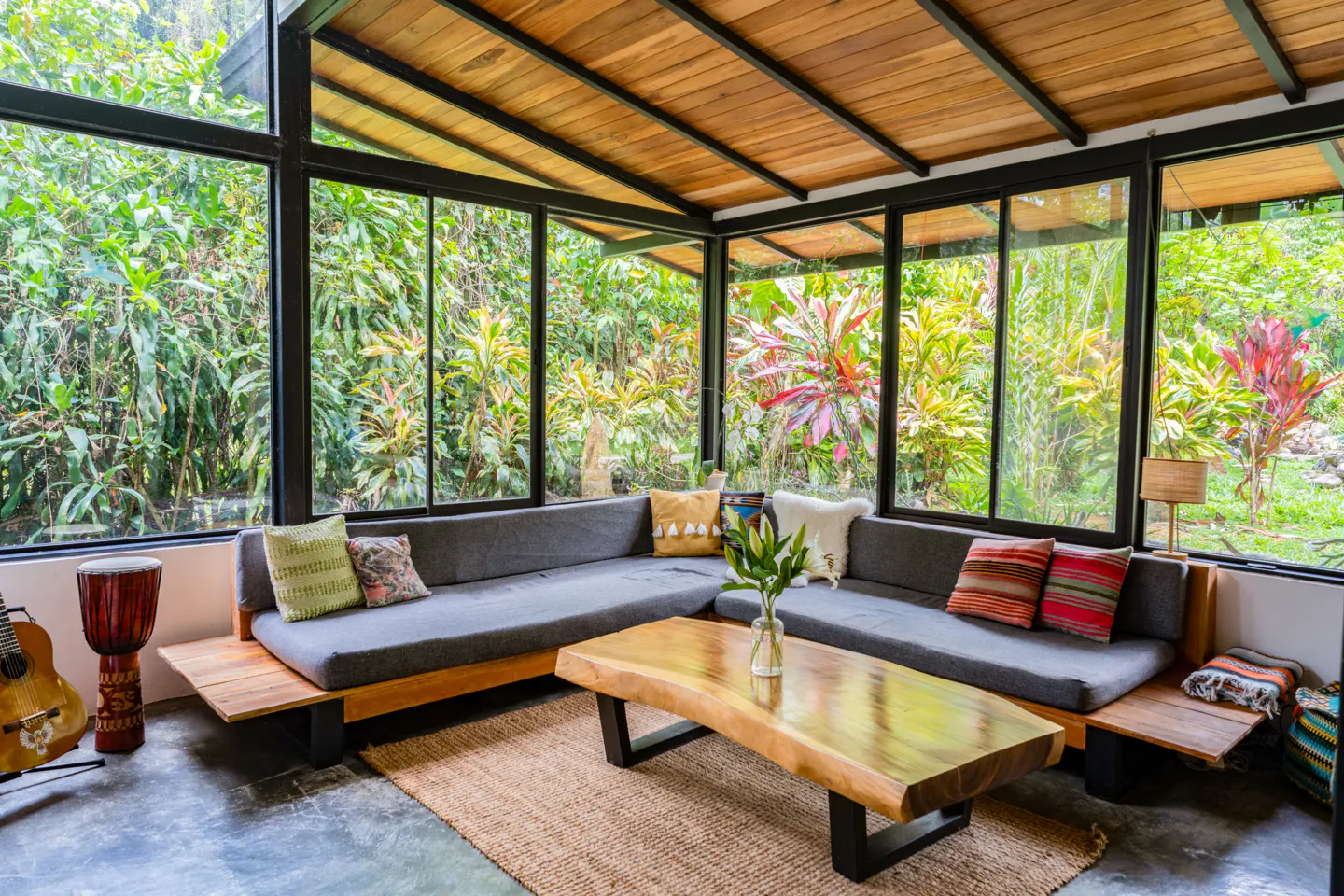 Bright sunroom with wood ceiling, black framed windows, and gray sectional sofa. Lush green foliage visible through the windows.