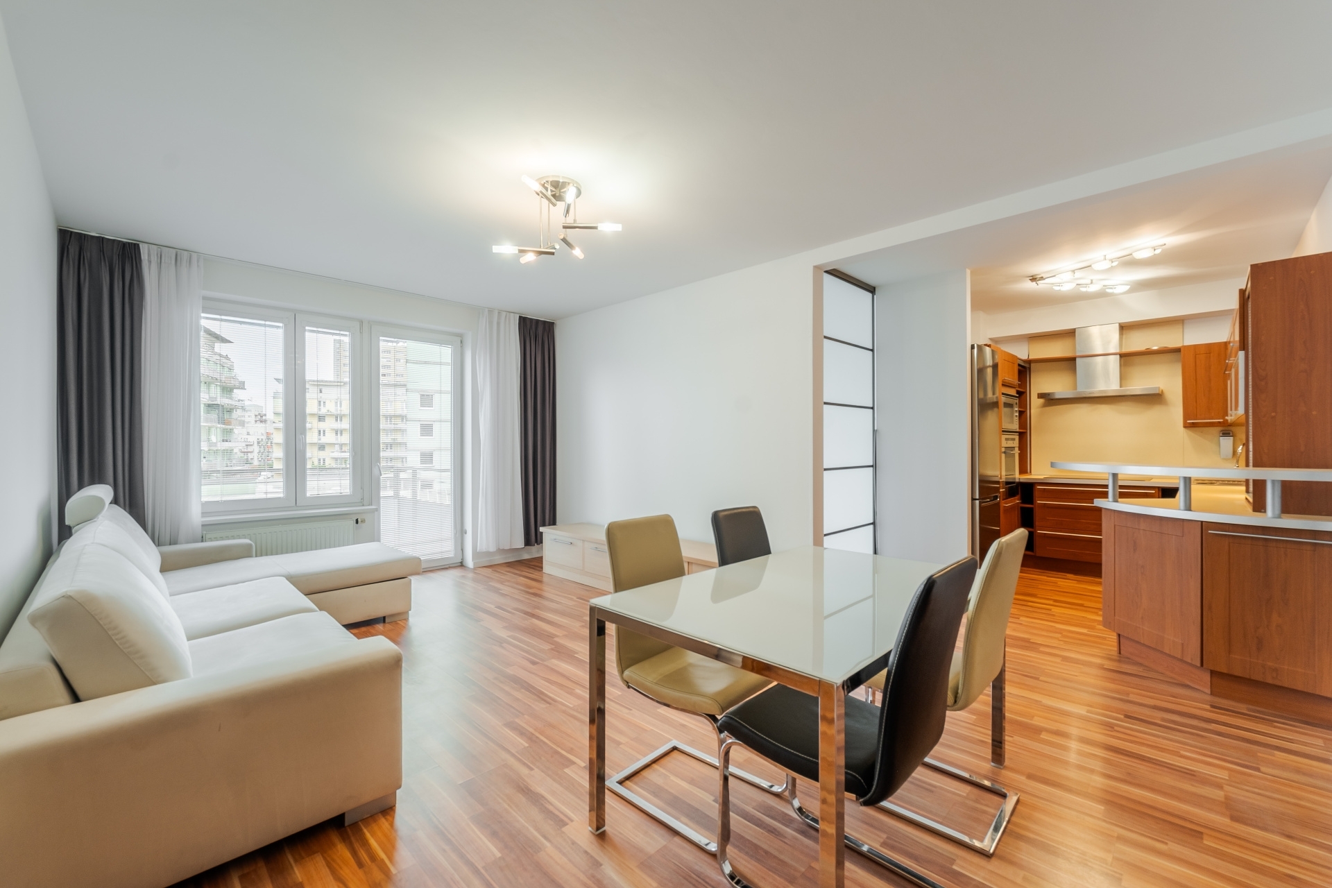 Open-plan living area with a white sofa, dining table with mixed chairs, and a wooden kitchen with stainless appliances.