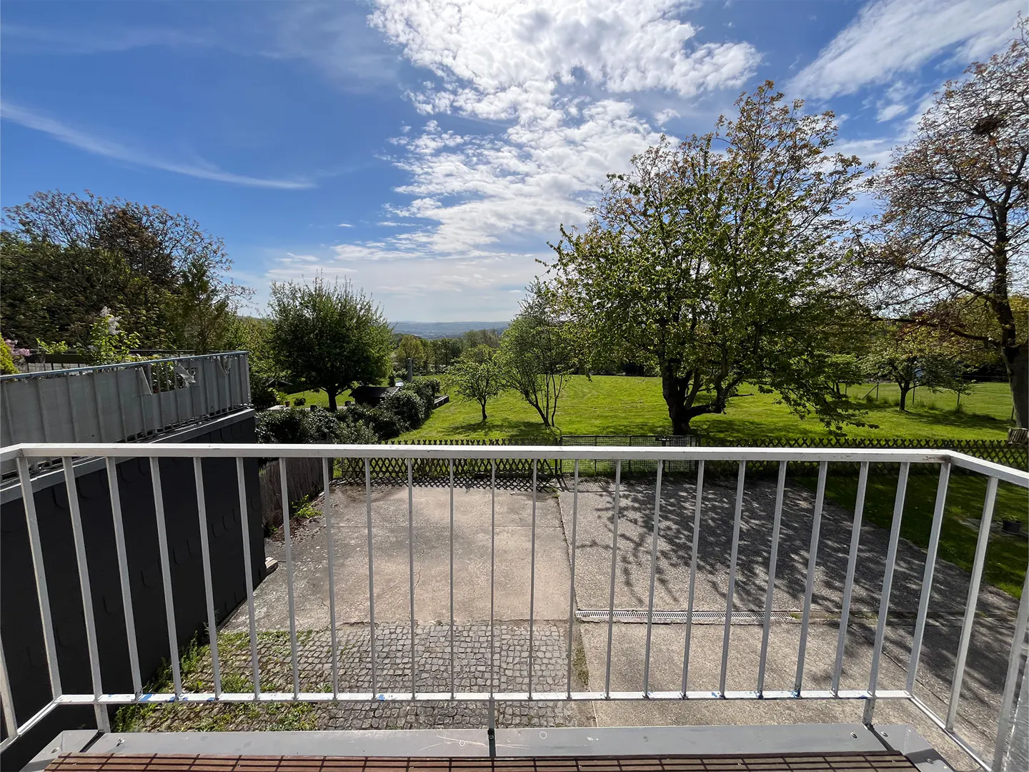 View from a balcony with a white railing overlooking a green lawn with trees under a blue sky with white clouds.