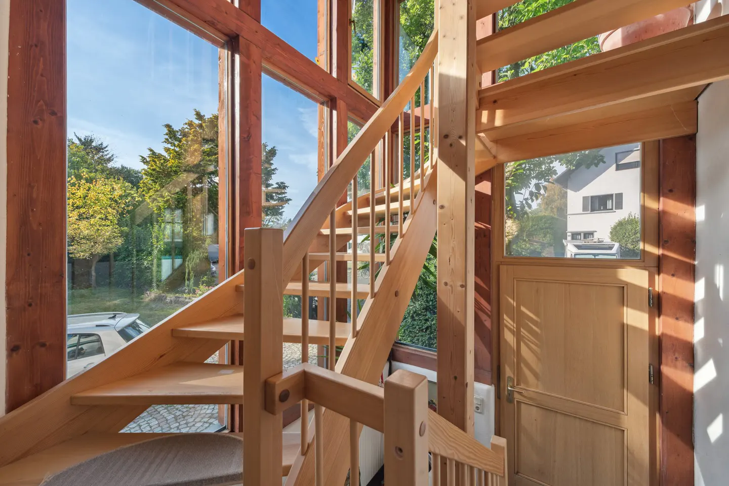 Interior view of a light wood staircase with a large window showing trees and a house. A wooden door is visible on the right.