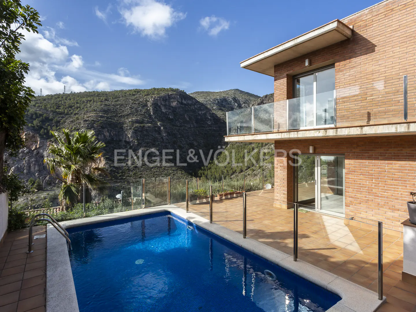 Modern brick house with a swimming pool, glass balcony, and a scenic mountain view under a bright blue sky.