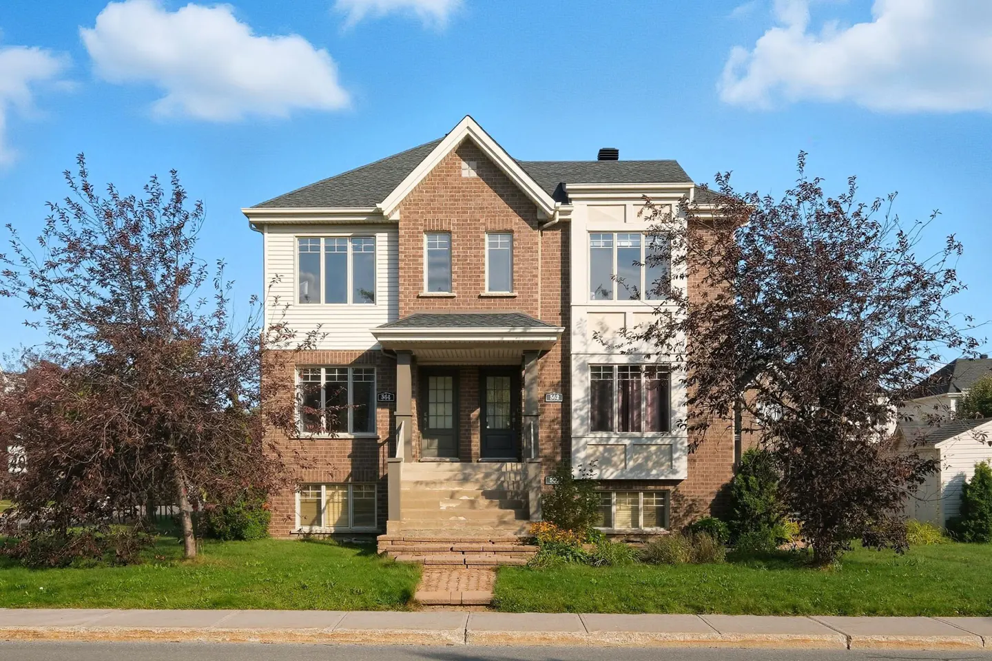 Two-story brick townhouse with a gray roof, white trim, and a small front yard with trees under a blue sky.