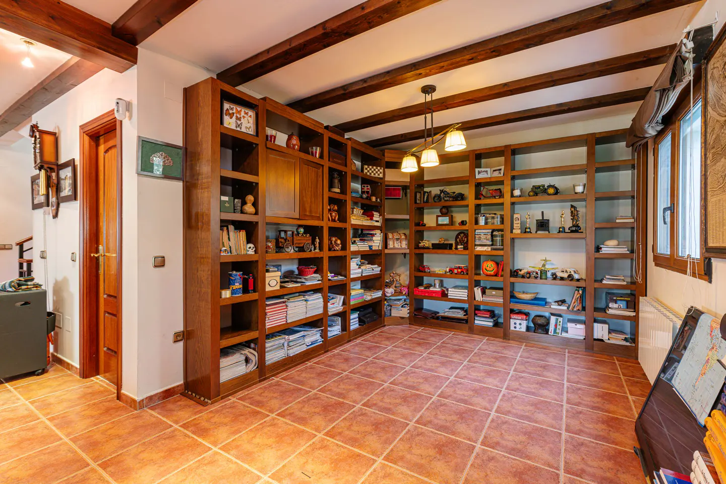 A room with a large, brown wooden bookcase filled with books and decorative objects. The floor is tiled in terracotta. Wooden beams are on the ceiling.