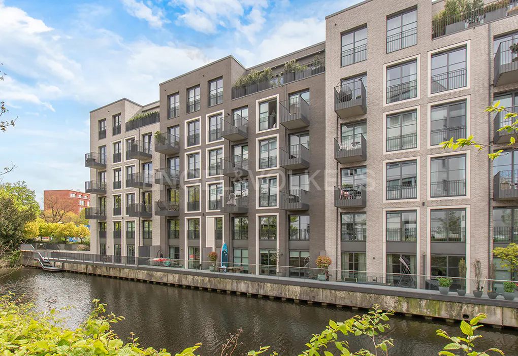 Modern apartment buildings with balconies line a canal under a cloudy sky. Green foliage in the foreground.