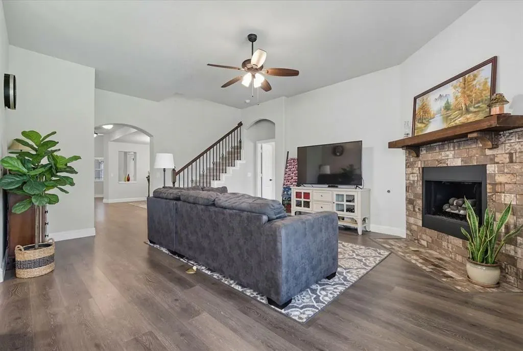 A living room with gray sofa, TV, fireplace, and wood floors. A ceiling fan and staircase are also visible.