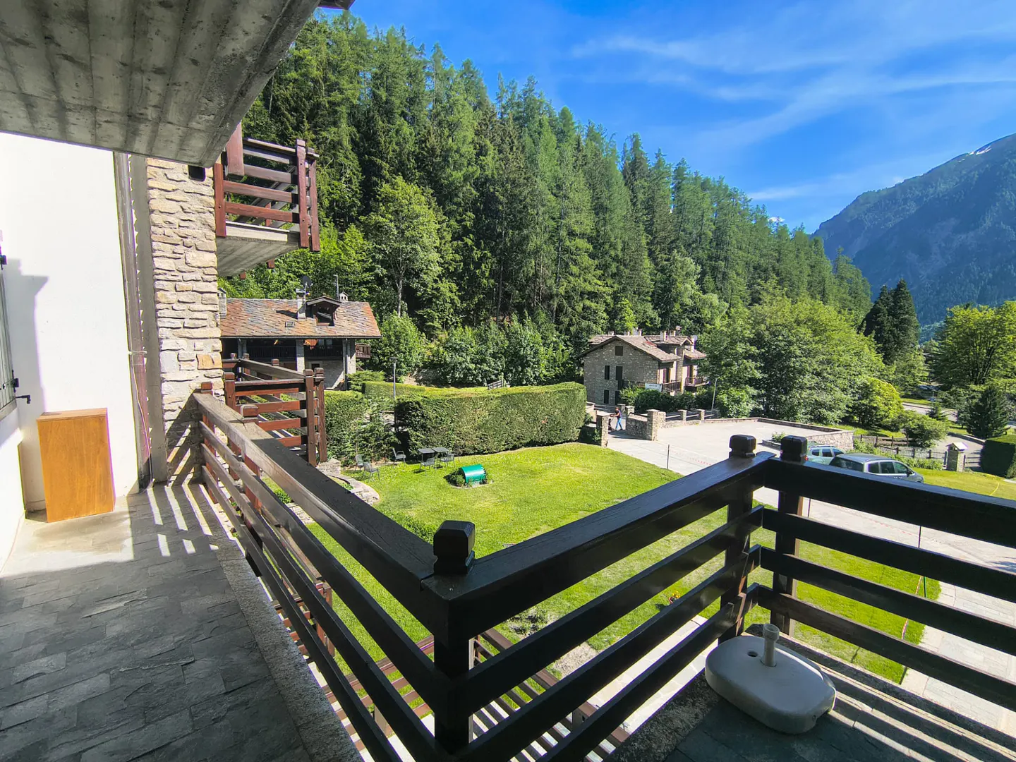 View from a stone balcony overlooking a green lawn, lush forest, and mountain landscape under a clear blue sky.