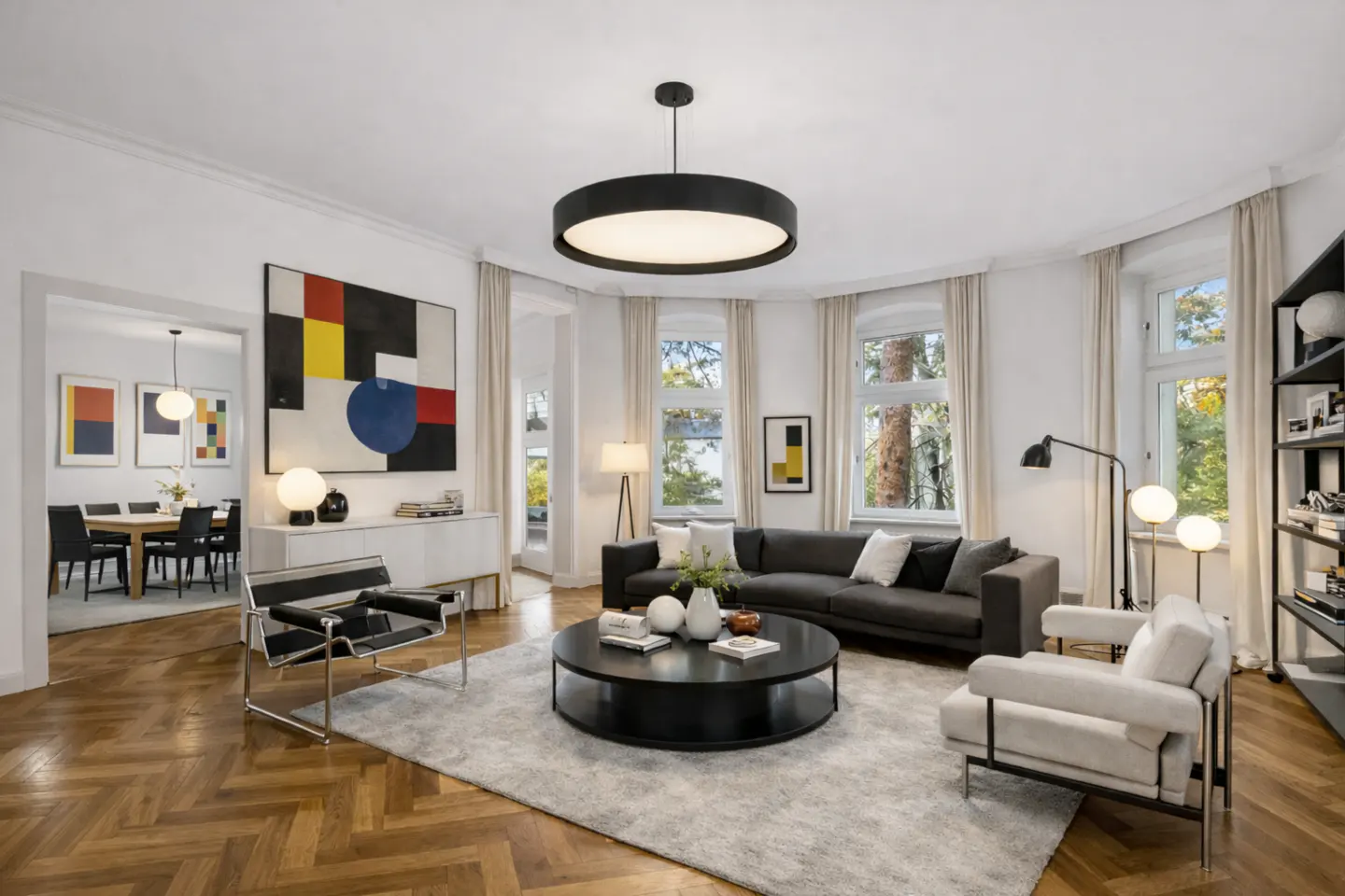 Bright living room with herringbone wood floors, gray sofa, round black table, and modern art. Large windows with white curtains.