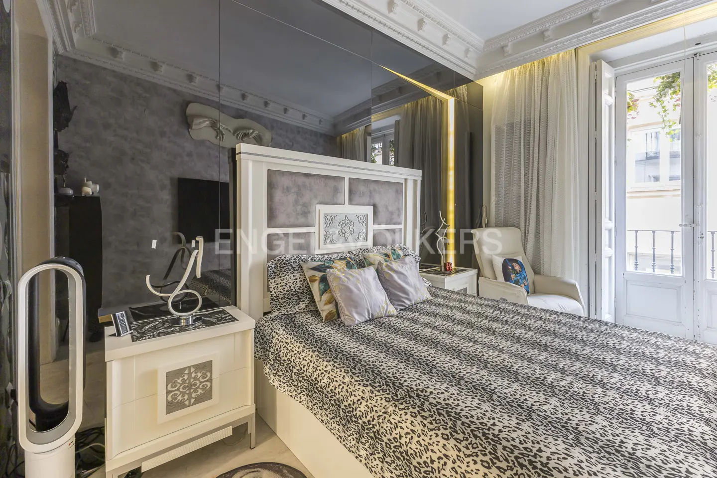 Bedroom with a large bed covered in a leopard-print blanket, white headboard, and mirrored wall. A white chair sits near a window with a balcony.