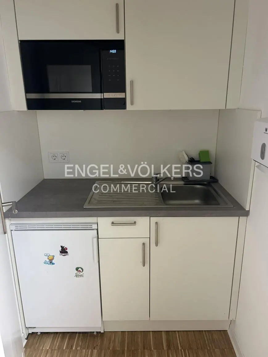A small kitchen area with white cabinets, a gray countertop, a stainless steel sink, and a black microwave. A small white refrigerator is on the left.