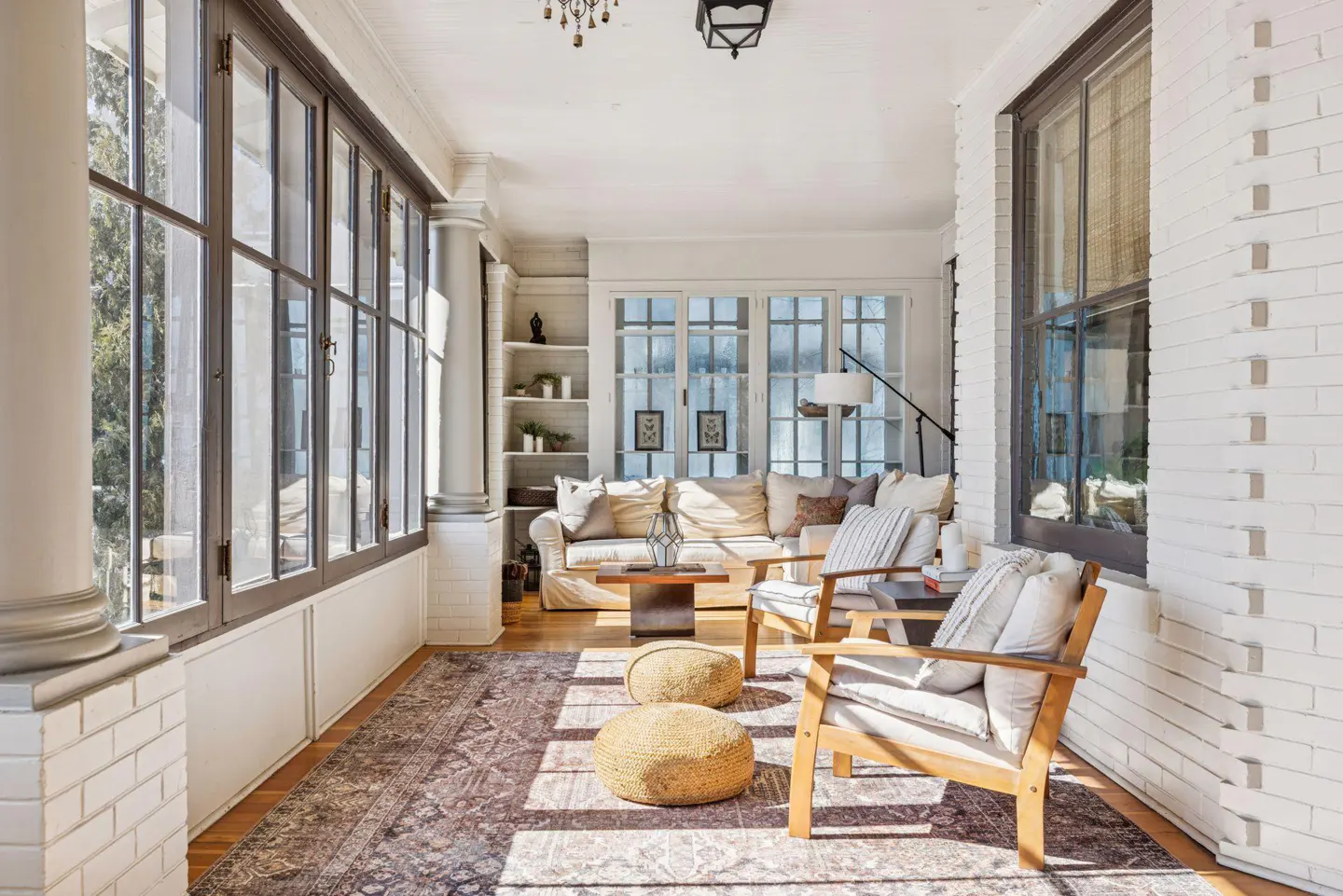 Bright sunroom with white brick walls, large windows, and a patterned rug. Two wooden chairs face a cream sofa with pillows.