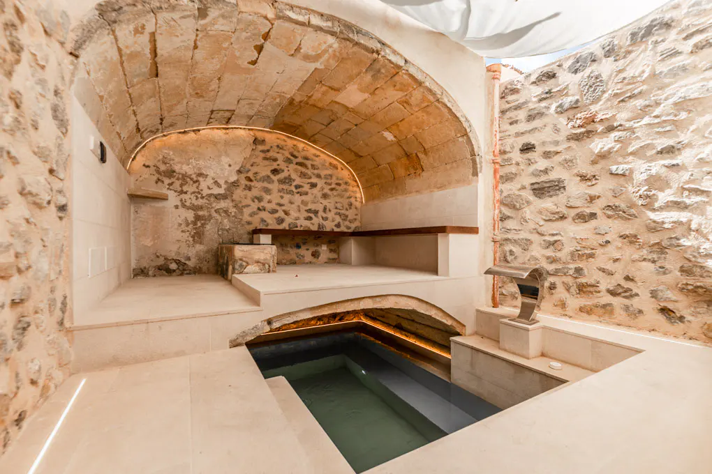Indoor pool area with stone walls and arched ceiling. A wooden bench sits in an alcove with warm lighting. The pool is rectangular and tiled.
