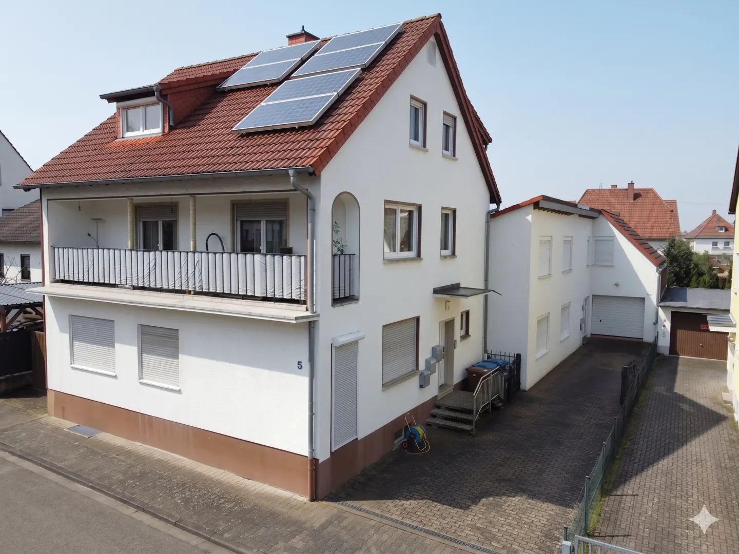 Two-story white house with a red tile roof and solar panels. A balcony is on the second floor. A driveway leads to a garage.
