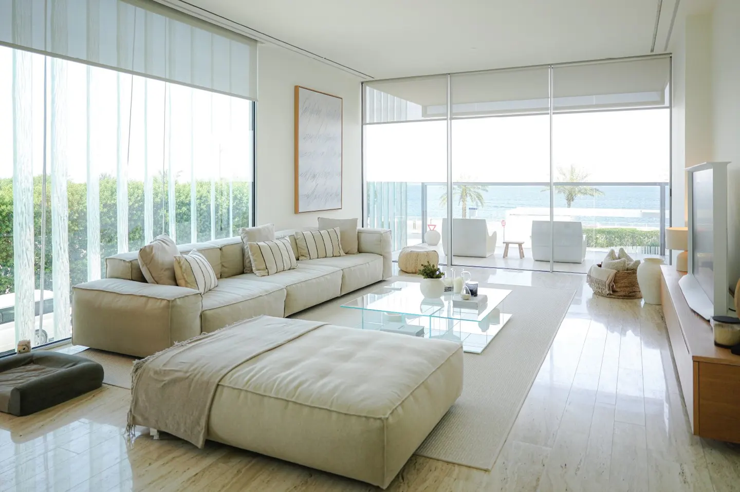 Bright, modern living room with a large beige sofa, glass coffee table, and ocean view through floor-to-ceiling windows.
