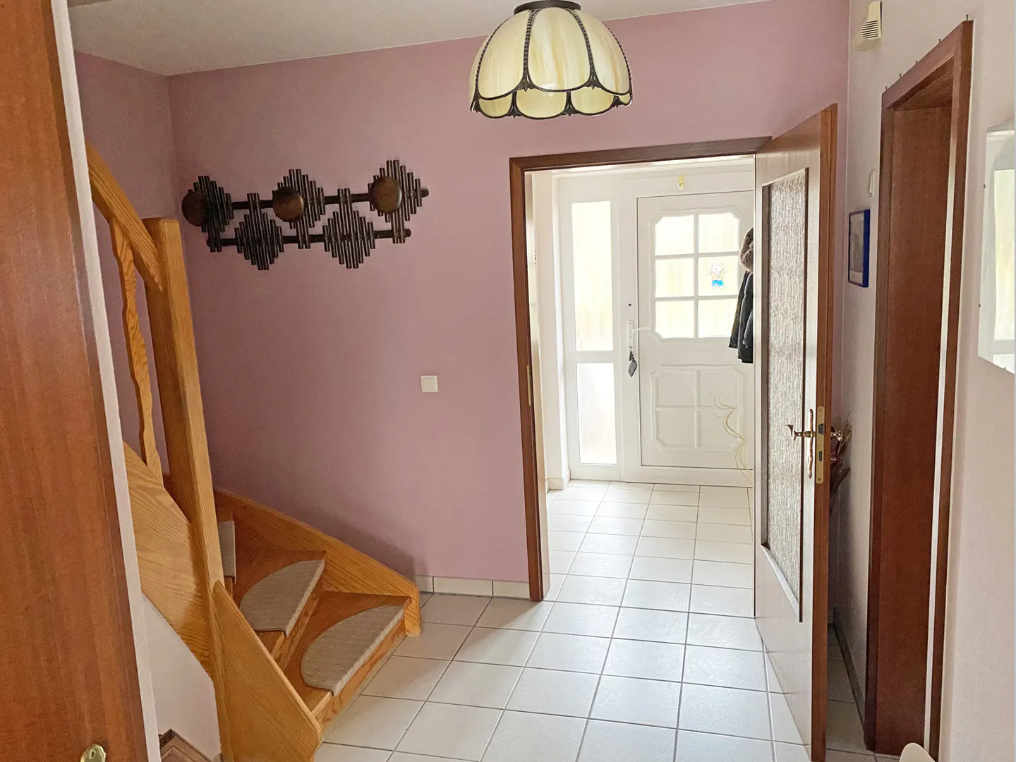 Interior view of a home's entryway with white tile floors, a wooden staircase, and a white front door. The walls are light purple.