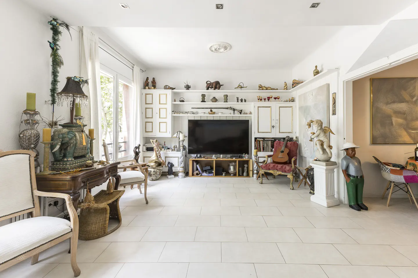 Bright, eclectic living room with white tile floor, built-in shelves, TV, and various decorative objects.