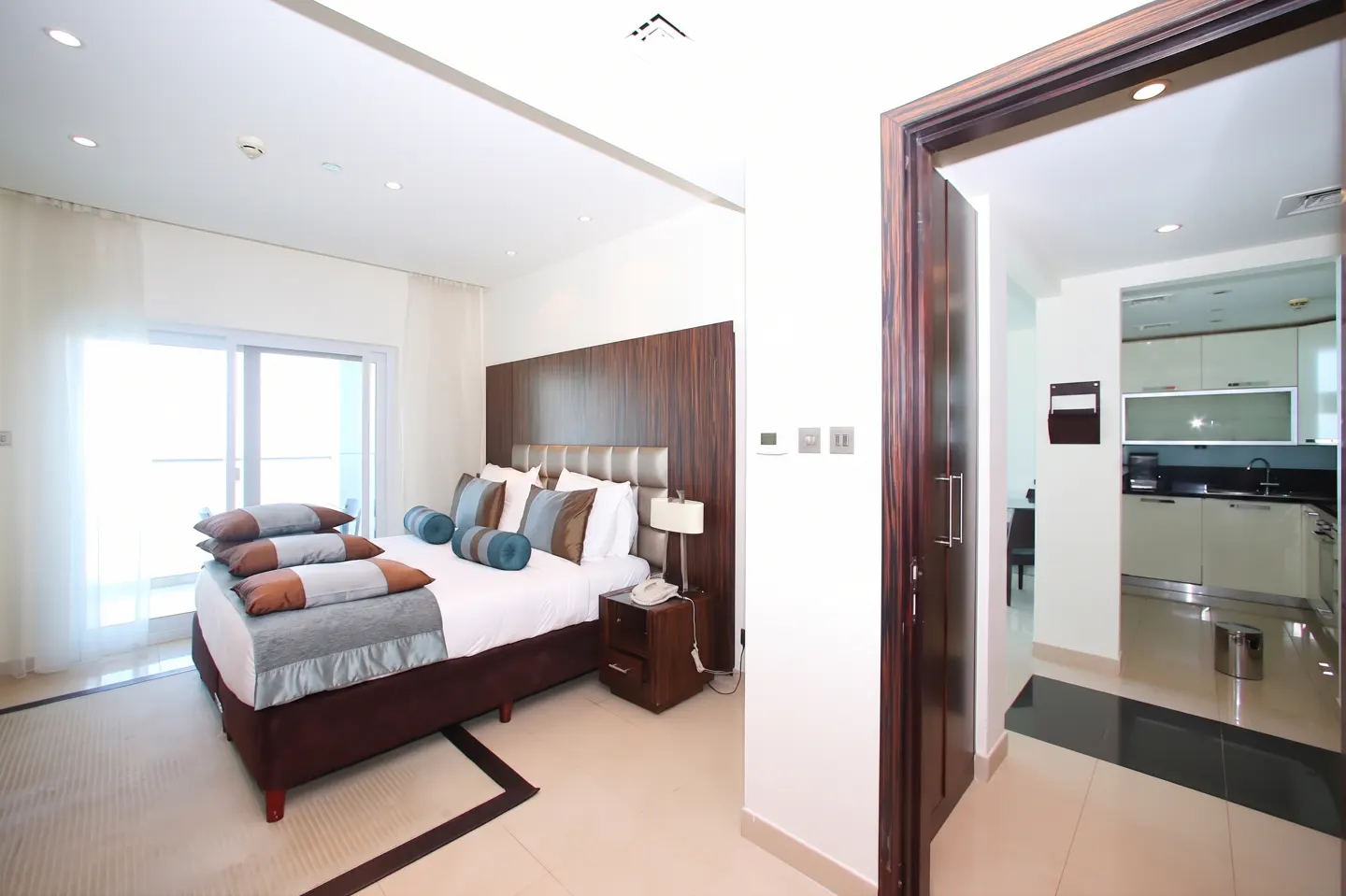A bright bedroom with a brown bed frame, white sheets, and blue pillows. A dark wood door leads to a kitchen with white cabinets.