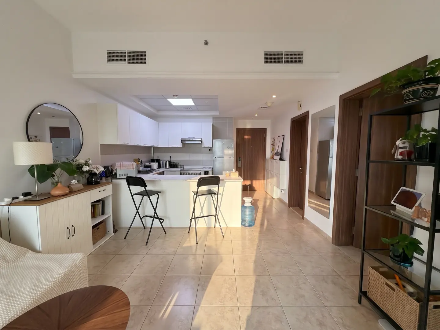 A bright, open-concept apartment with a white kitchen, bar stools, and a living area with a round mirror and shelving unit.
