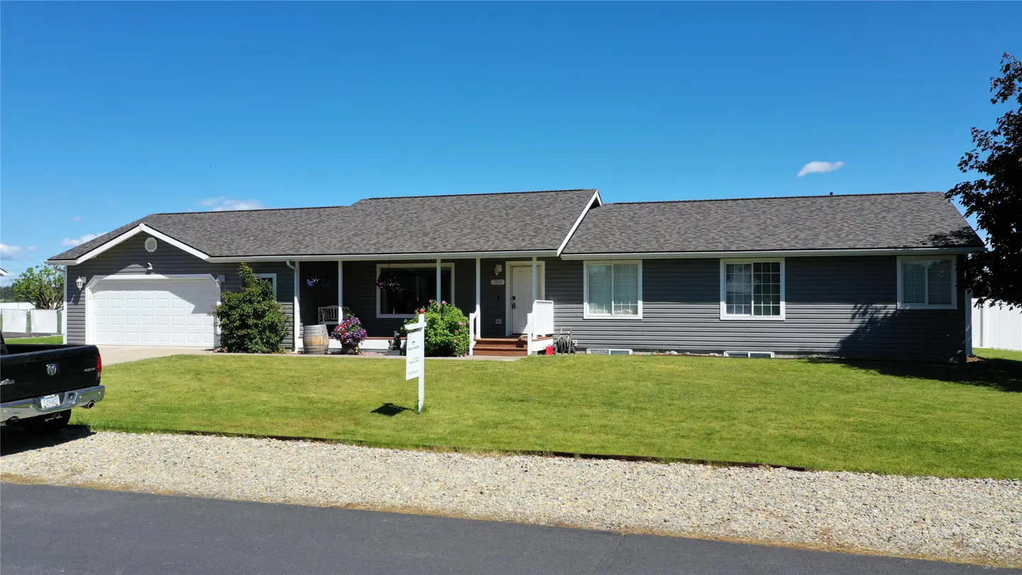 A gray single-story house with a white garage door and a "For Sale" sign on a green lawn.