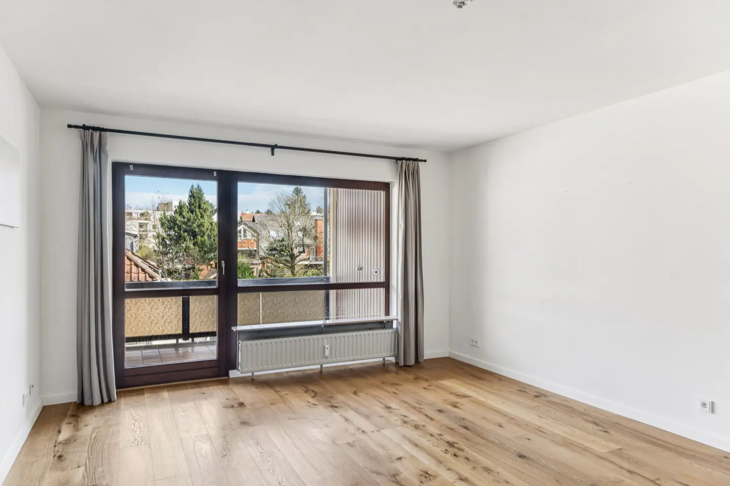 Bright, empty room with wood floors, white walls, and a large window/door to a balcony with a city view. Gray curtains frame the window.