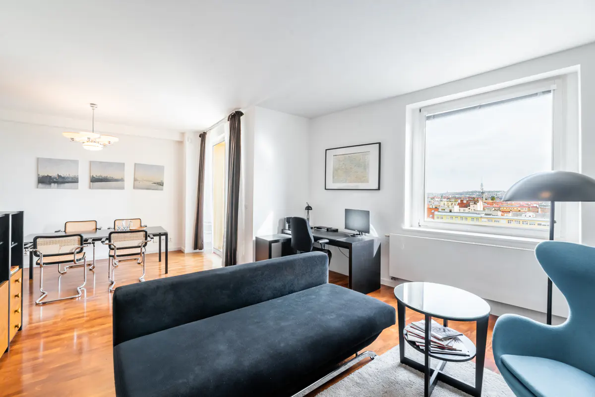 Bright living room with hardwood floors, a black sofa, a desk, and a dining table with four chairs. A large window overlooks a cityscape.